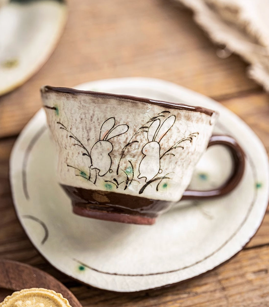 Handmade ceramic mug with rabbit design and coaster, cream and brown, on wooden table.